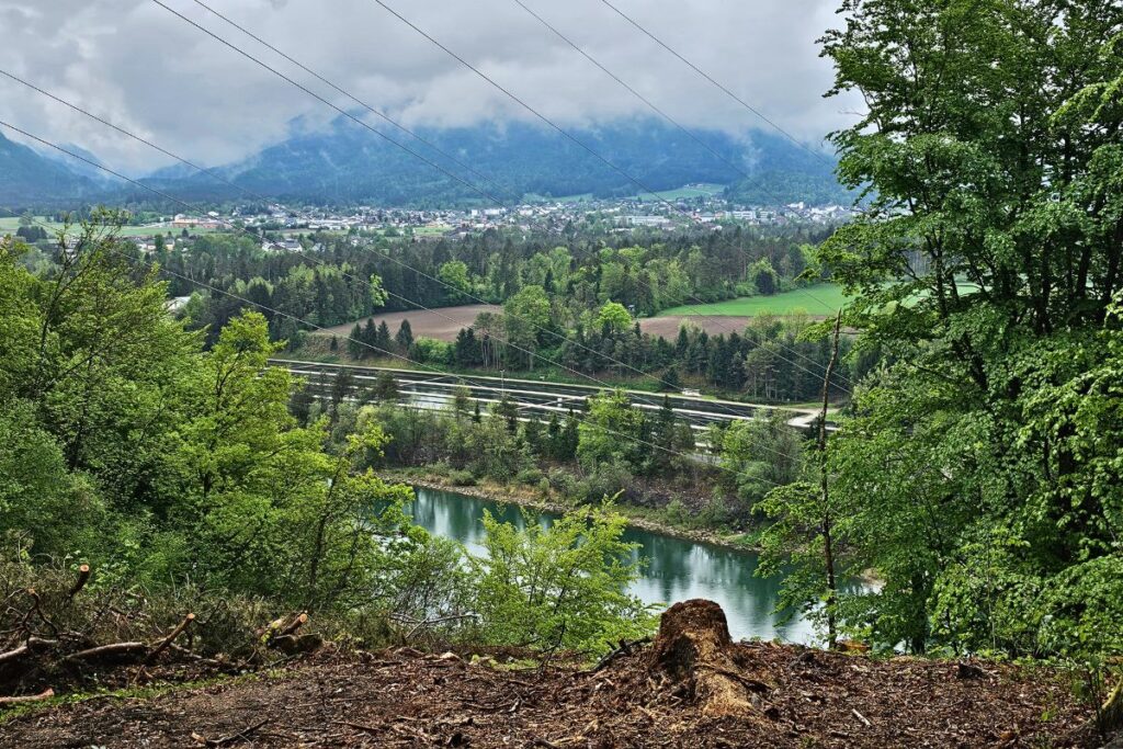 Ein weiter Panoramablick von einem erhöhten Aussichtspunkt im Rosental hinab auf den langgestreckten, bläulichen Ferlacher Stausee, eingebettet in dichte grüne Wälder und Berge.