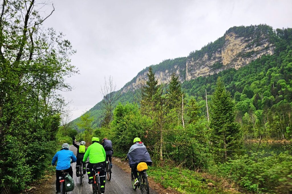 Mehrere Radfahrer befahren hintereinander den flachen, asphaltierten Drauradweg direkt neben dem breiten, ruhig dahinfließenden Fluss im strahlenden Sonnenschein.