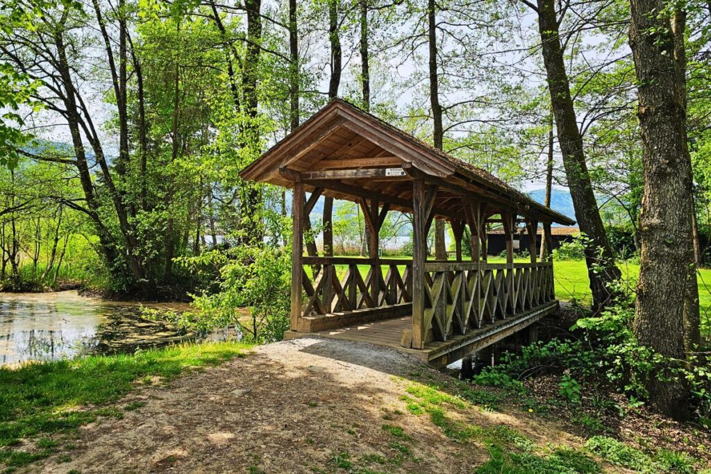 Eine idyllische, überdachte Holzbrücke inmitten von saftig grünen Bäumen an einem kleinen Bachlauf auf dem Radweg Richtung Keutschacher See.