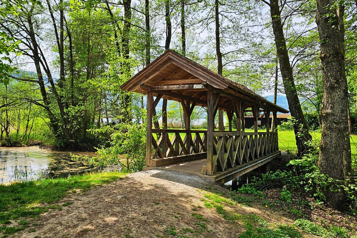 Eine idyllische, überdachte Holzbrücke inmitten von saftig grünen Bäumen an einem kleinen Bachlauf auf dem Radweg Richtung Keutschacher See.