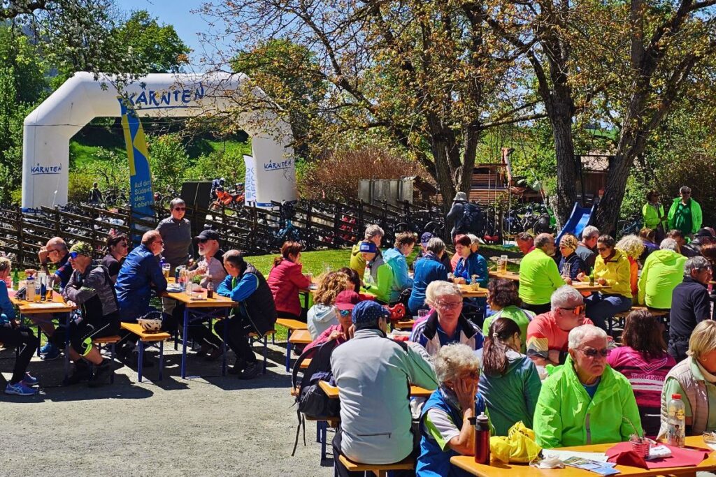 Zahlreiche Radfahrer sitzen in der Sonne an hölzernen Tischen im Freien bei einem organisierten Mittagsstopp an der Buschenschenke Brunnerhof unter schattigen Bäumen in Kärnten.