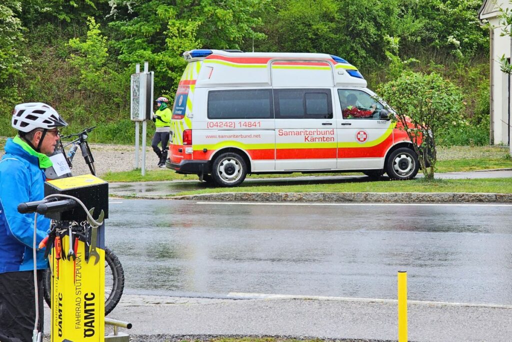 Ein weiß-roter Rettungswagen (RTW) des Samariterbundes Kärnten steht auf nasser Straße an einem gelben ÖAMTC-Fahrrad-Stützpunkt.