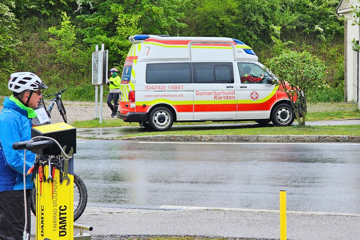 Ein weiß-roter Rettungswagen (RTW) des Samariterbundes Kärnten steht auf nasser Straße an einem gelben ÖAMTC-Fahrrad-Stützpunkt.