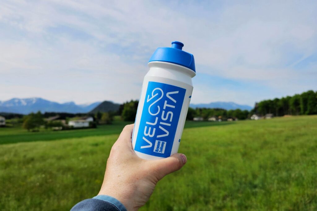 Eine Hand hält eine weiße Trinkflasche mit blauem „VELOVISTA“-Schriftzug vor einer grünen Wiese mit den Kärntner Alpen im Hintergrund.
