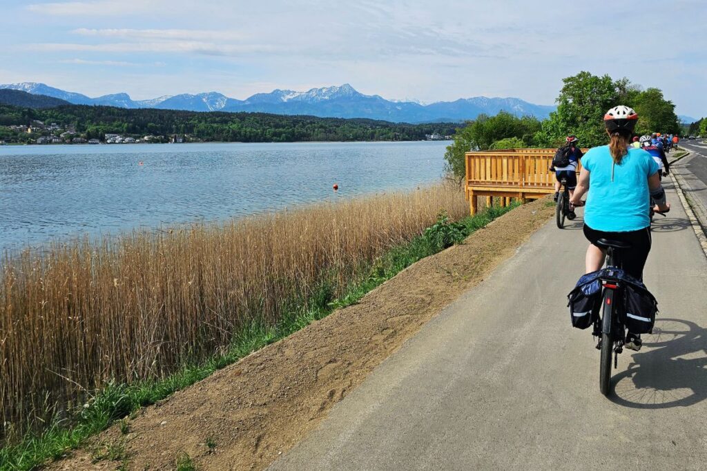 Eine Gruppe von Radfahrern fährt an einem sonnigen Tag auf einem flachen Asphaltweg direkt am glitzernden, türkisblauen Ufer des Wörthersees entlang.