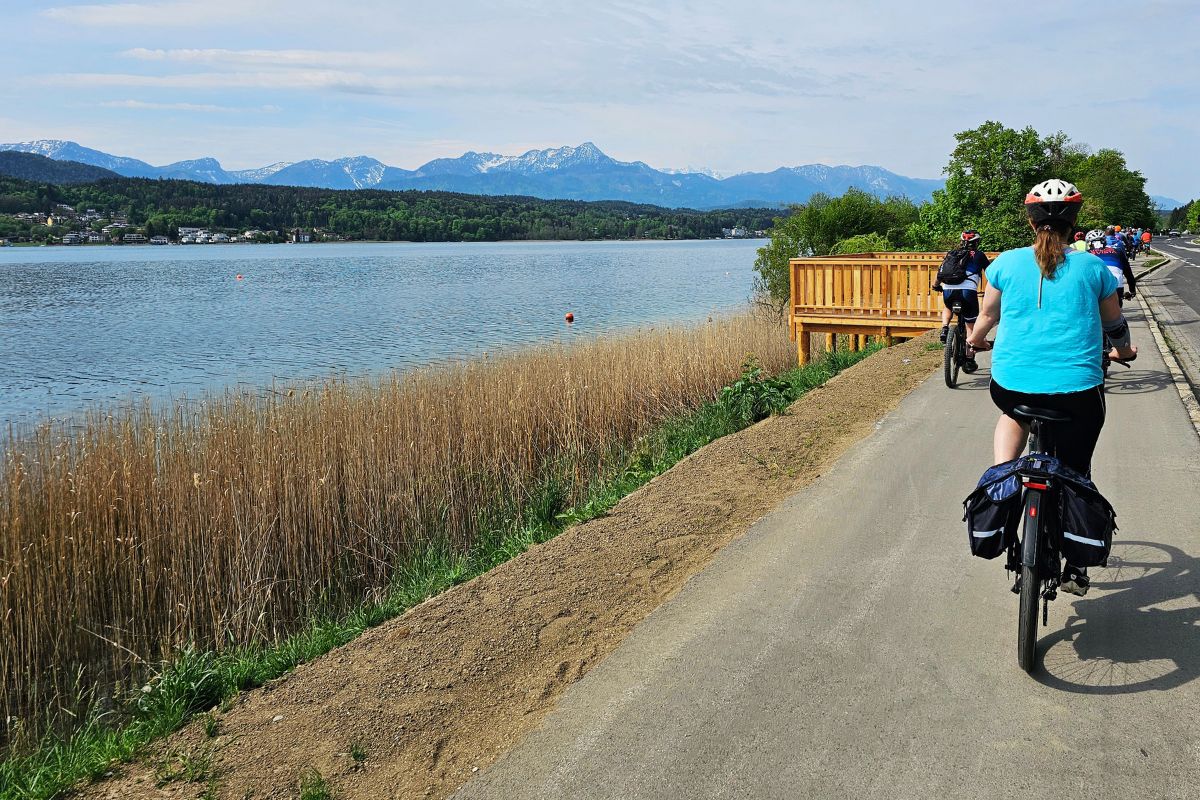 Eine Gruppe von Radfahrern fährt an einem sonnigen Tag auf einem flachen Asphaltweg direkt am glitzernden, türkisblauen Ufer des Wörthersees entlang.