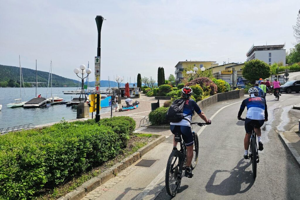 Die Velovista-Tourguides und Radfahrer posieren gut gelaunt in sportlicher Radkleidung für ein Gruppenbild an einem Holzgeländer direkt vor der Kulisse des Wörthersees.