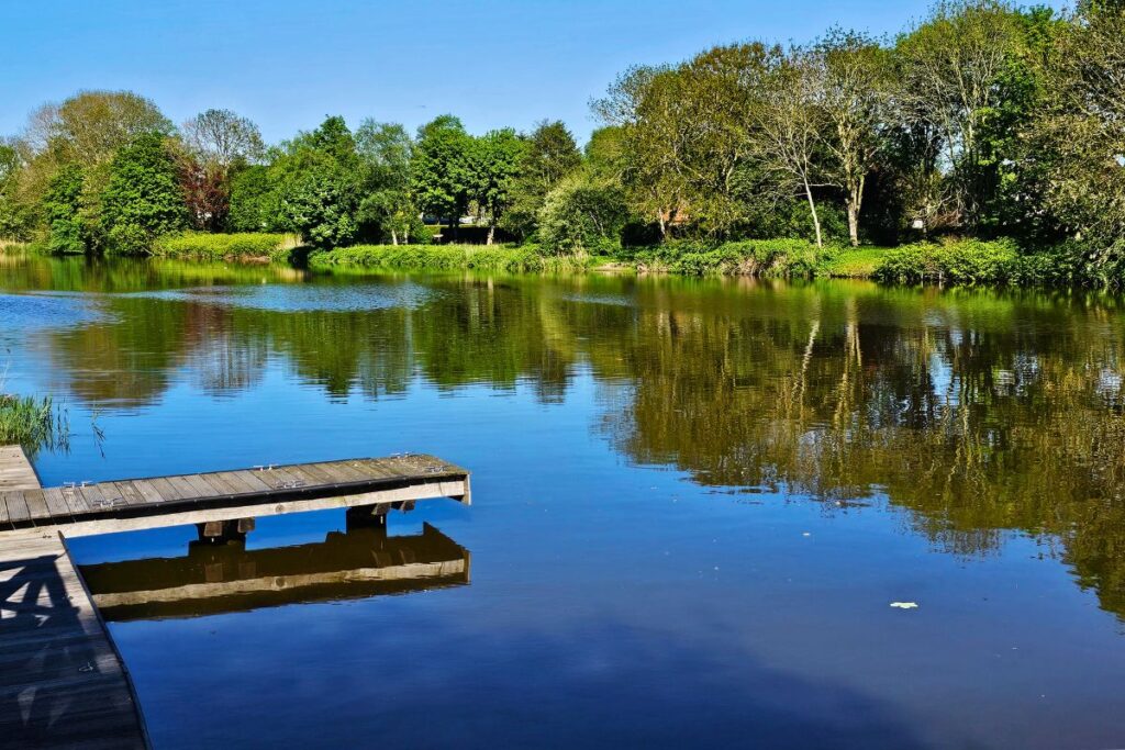 Ein hölzerner Badesteg ragt in das spiegelglatte, blaue Wasser der Braake in Brunsbüttel, umgeben von dichten grünen Bäumen und Spiegelungen.