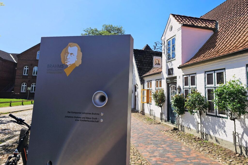 Eine graue Infotafel der Brahms-Gesellschaft mit dem gezeichneten Porträt von Johannes Brahms im Vordergrund, dahinter führt ein Ziegelweg zu einem weißen historischen Wohnhaus.