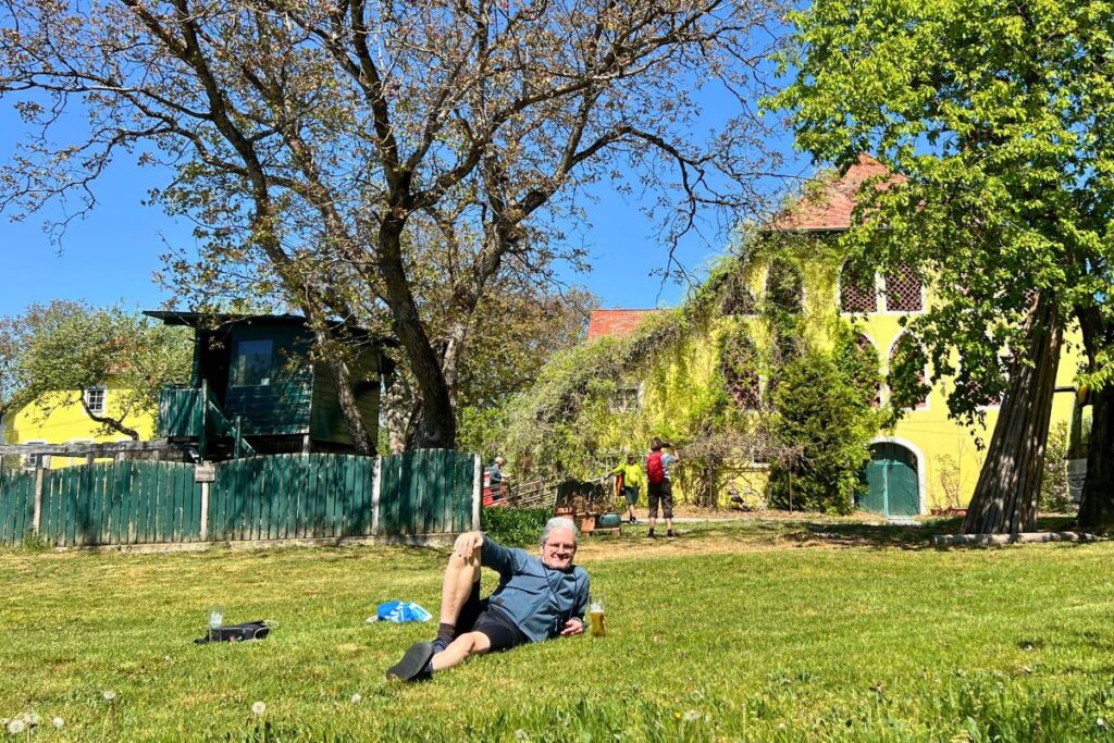 Nicolo Martin liegt entspannt im grünen Gras auf der Wiese vor der gelben, mit Kletterpflanzen bewachsenen Fassade der Buschenschenke Brunnerhof.