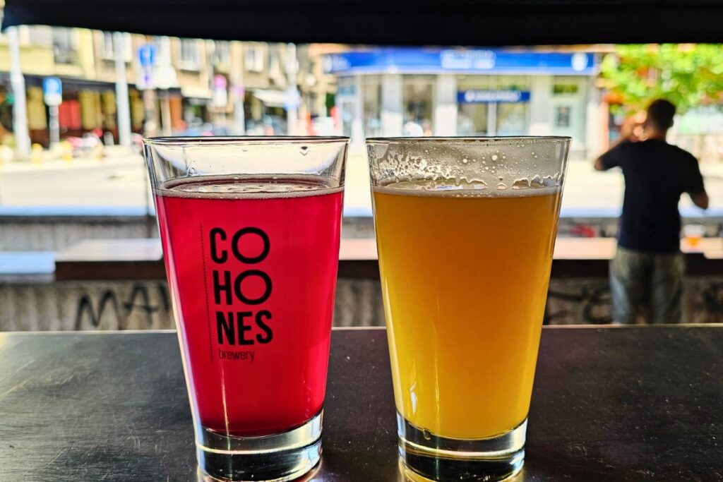 Zwei Gläser Craft Beer stehen auf einem Tresen vor einem Fenster zur Straße; links ein tiefrotes Bier im Cohones-Brewery-Glas, rechts ein trübes gelbes Hazy IPA.
