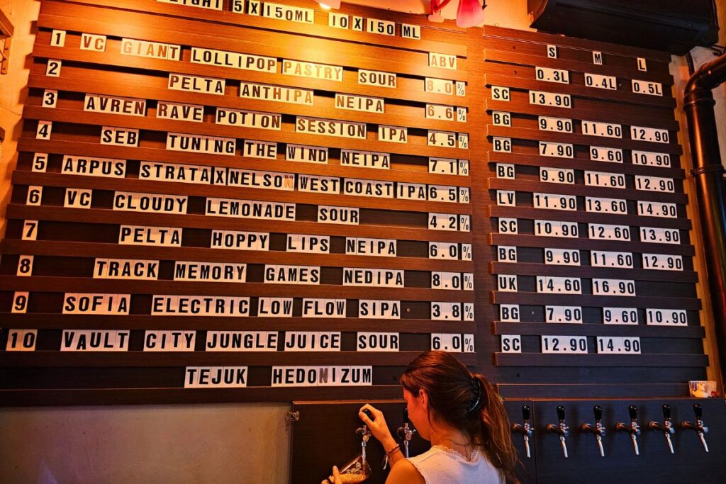 Eine große hölzerne Stecktafel mit der aktuellen Bierauswahl einer Craft-Beer-Bar in Sofia, während eine Barkeeperin im Vordergrund frisches Bier zapft.