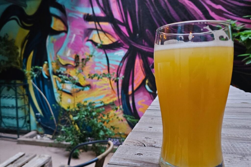Ein großes Glas hellgelbes, trübes Craft Beer steht im Vordergrund auf einem Holztisch vor einer farbenfrohen Streetart-Wandmalerei mit dem Gesicht einer Frau.