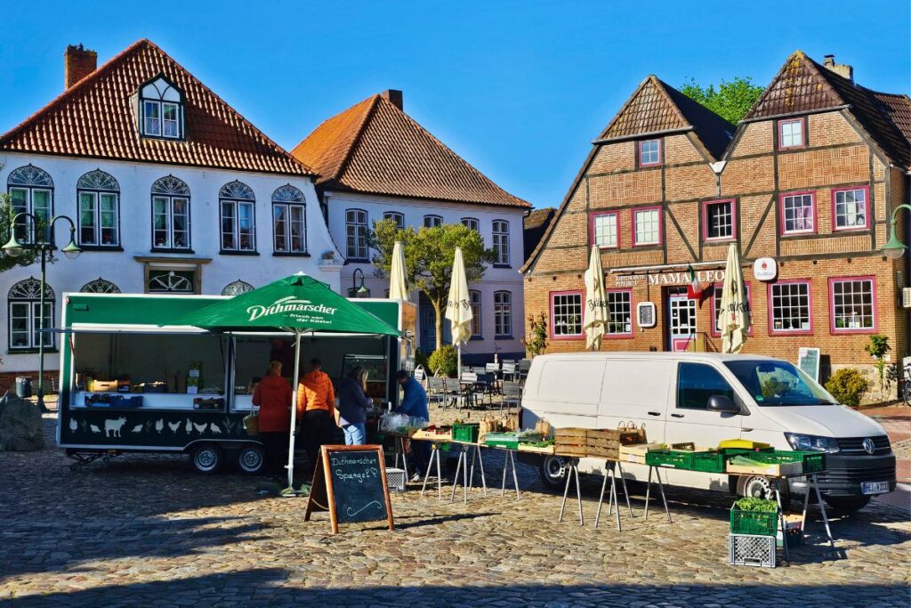 Ein sonniger Wochenmarkt auf dem historischen Kopfsteinpflaster vor bunten Giebel- und Fachwerkhäusern in Meldorf, Dithmarschen, mit einem Marktstand für frischen regionalen Spargel.