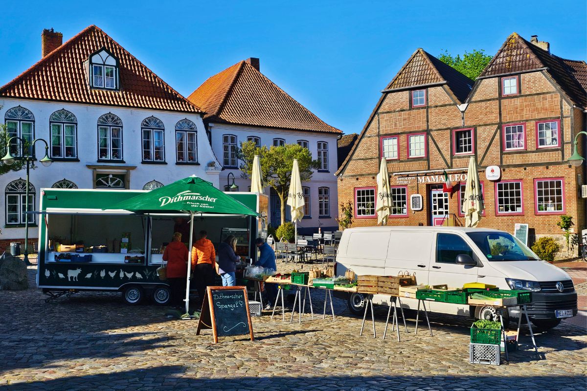 Ein sonniger Wochenmarkt auf dem historischen Kopfsteinpflaster vor bunten Giebel- und Fachwerkhäusern in Meldorf, Dithmarschen, mit einem Marktstand für frischen regionalen Spargel.