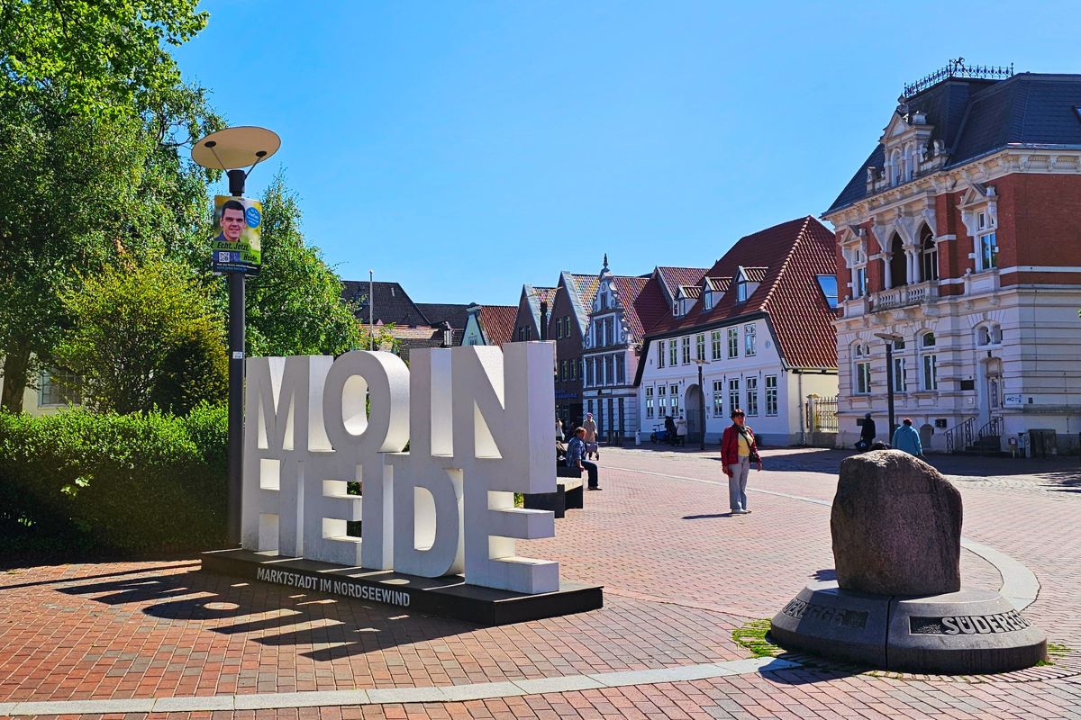 Ein großer, weißer dreidimensionaler Schriftzug mit der Aufschrift "MOIN HEIDE" steht auf dem gepflasterten Südermarkt vor historischen Giebelhäusern. Rechts im Vordergrund ist ein runder Gedenkstein platziert.