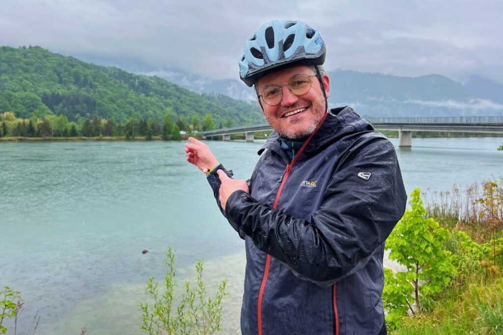 Nicolo Martin im Regen in einer dunklen Outdoorjacke und Fahrradhelm lächelt und zeigt auf die breite Drau und eine Brücke im Hintergrund.