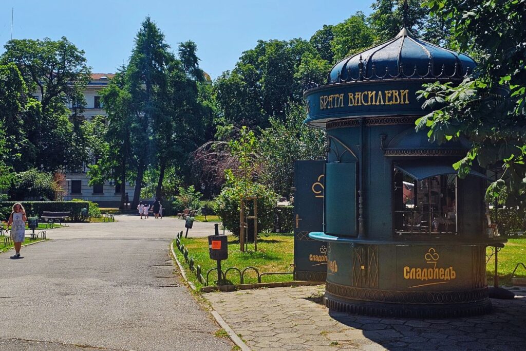Ein historischer, dunkelgrüner Eispavillon mit goldener kyrillischer Aufschrift der Gebrüder Vassilevi in einem sonnigen Park in Sofia.