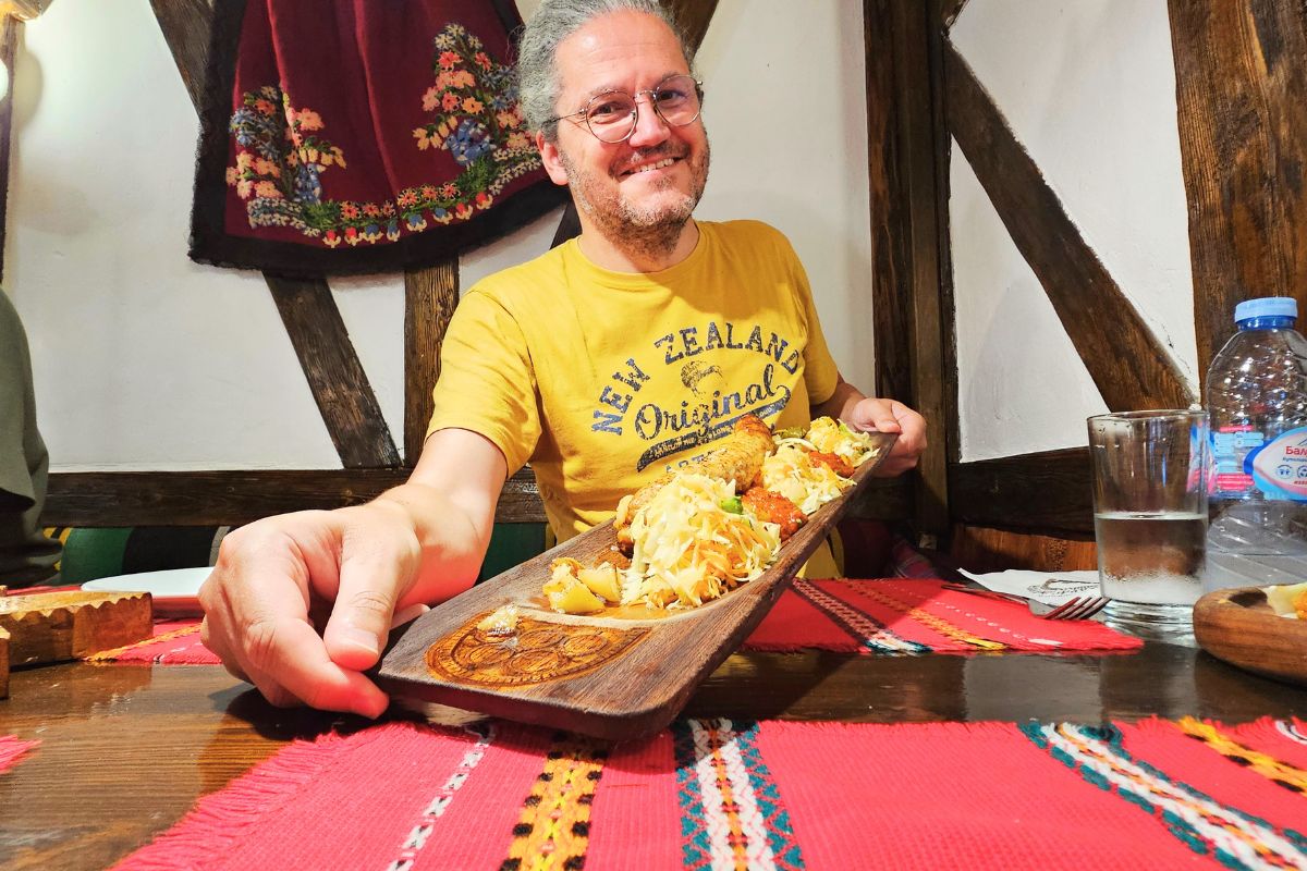 Reiseblogger Nicolo Martin präsentiert lächelnd eine lange Holzplatte mit traditionellen bulgarischen Grillspezialitäten in einem Restaurant in Sofia.