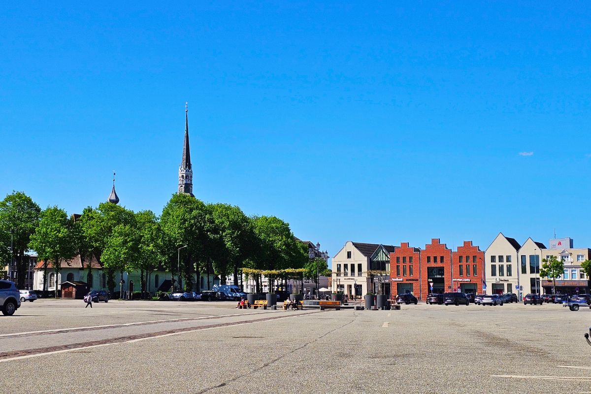 Der weitläufige, unbebaute und gepflasterte Marktplatz in Heide unter strahlend blauem Himmel, umgeben von grünen Alleen und modernen sowie historischen Gebäudereihen.