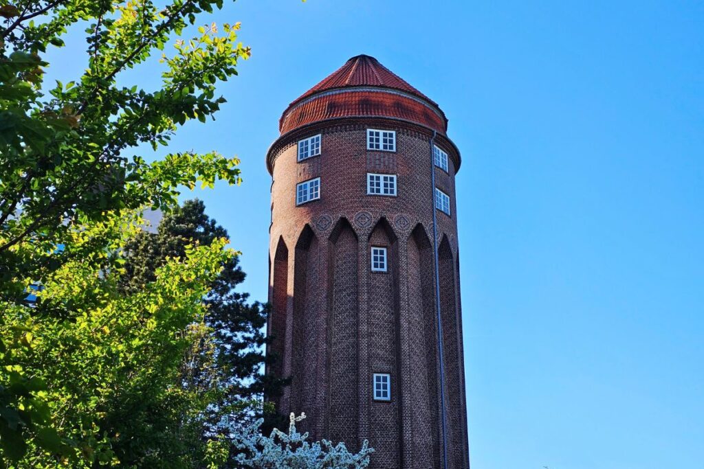 Der imposante historische Wasserturm aus rotem Backstein in Brunsbüttel ragt mit seinem runden, ziegelgedeckten Dach hinter grünen Bäumen empor.