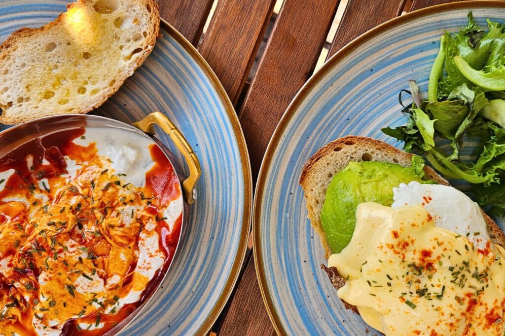 Zwei stilvolle blau-gestreifte Teller mit pochierten Eiern auf Avocado-Toast und Panagyurski Eggs im Pfännchen bei Izzy's Coffee in Sofia von oben fotografiert.