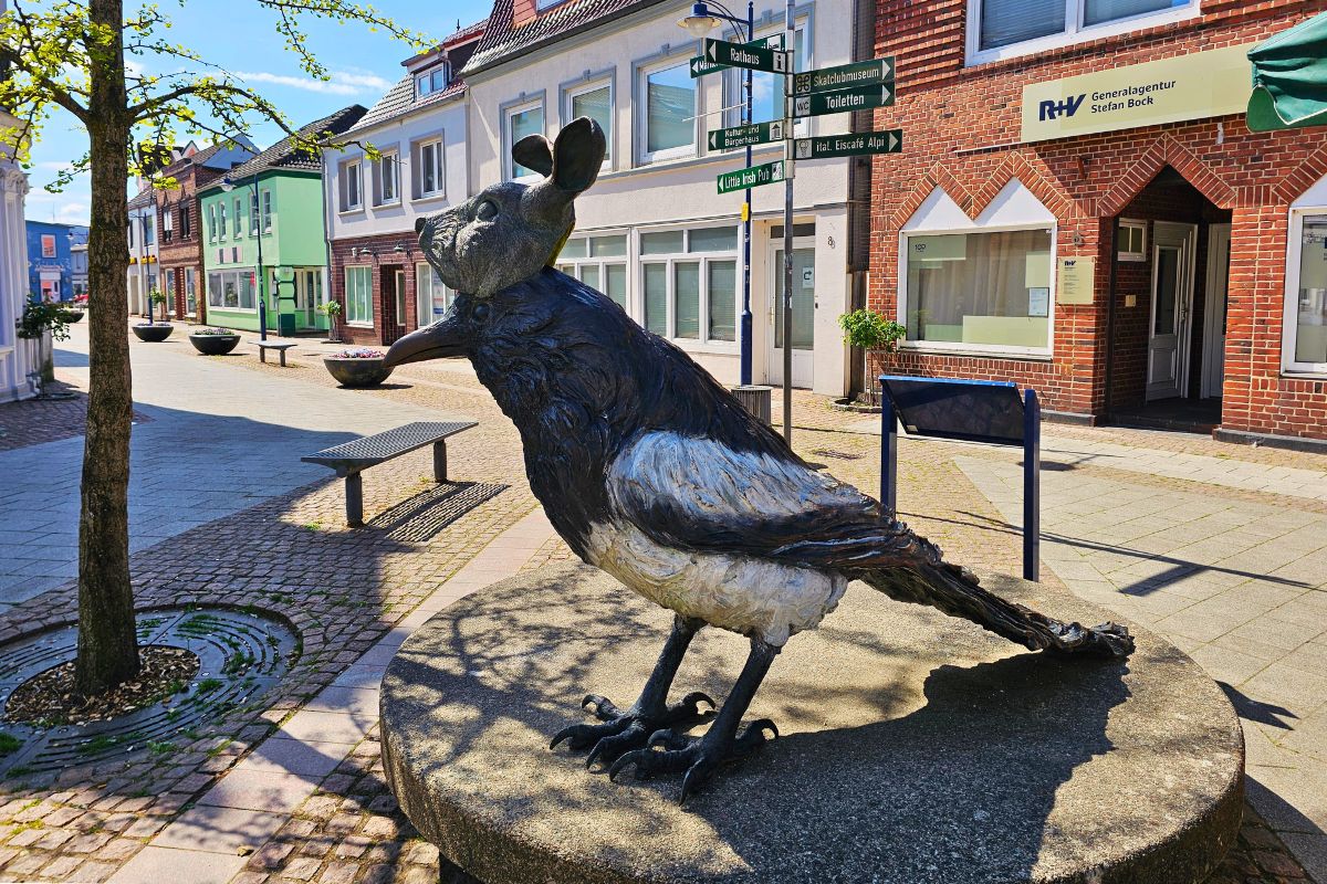 Eine lebensgroße Skulptur einer Elster, die eine Hasenmaske auf dem Kopf trägt, auf einem runden Steinsockel in der Fußgängerzone von Marne.