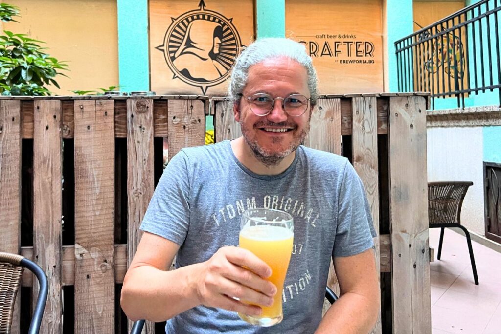 Nicolo Martin sits lächelnd im Außenbereich der Crafter Bar in Sofia und hält ein Glas naturtrübes Craft Beer in der Hand.