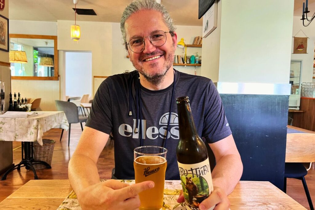 Nicolo Martin sitzt gut gelaunt an einem Tisch in einem Lokal, zeigt auf ein Glas Bier und hält eine Flasche Divo Pivo.