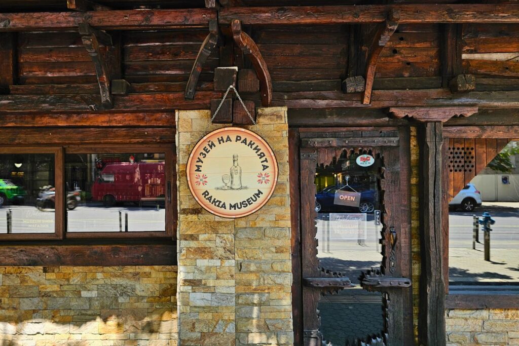 Der rustikale Holz- und Steineingang des Rakia Museums in Sofia mit einem runden Holzschild mit kyrillischer und englischer Aufschrift.
