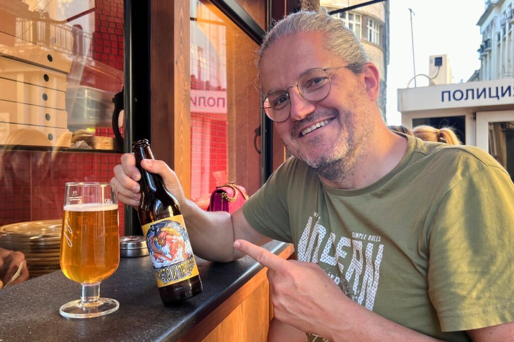 Reiseblogger Nicolo Martin lächelt in die Kamera und zeigt auf eine Bierflasche der bulgarischen Blek Pine Brewery mit buntem Etikett.