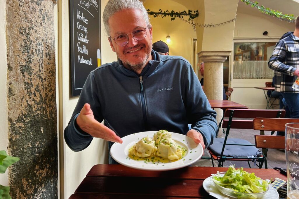 Reiseblogger Nicolo Martin sitzt lächelnd an einem Holztisch im Gasthaus und präsentiert einen Teller mit drei Kärntner Kasnudeln, daneben steht ein frischer grüner Salat.