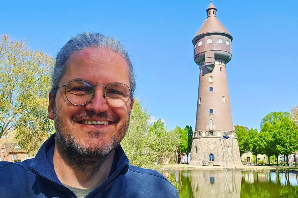 Reiseblogger Nicolo Martin lächelt im Vordergrund eines Selfies in die Kamera, im Hintergrund thront der historische Heider Wasserturm hinter einem kleinen Teich unter klarem blauem Himmel.