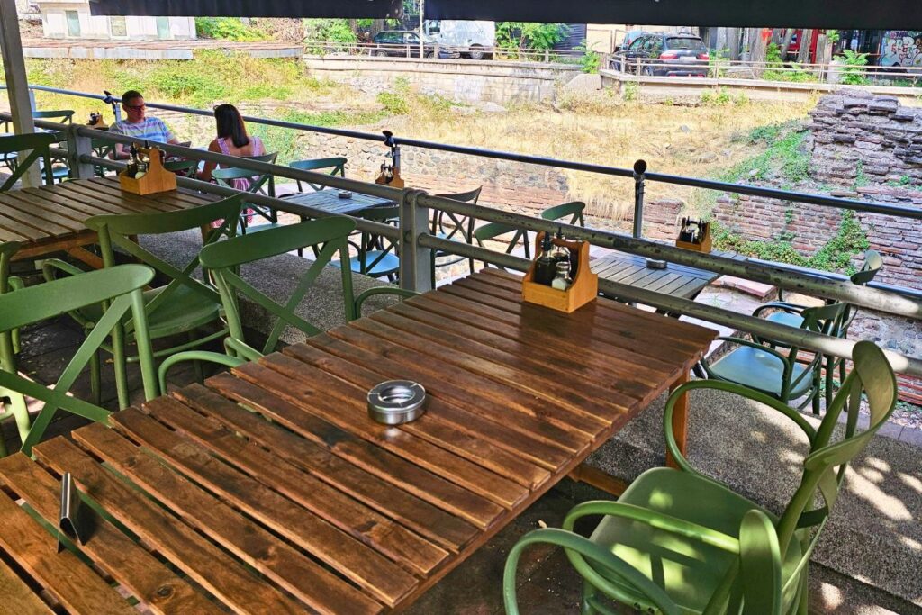 Die einladende Holzterrasse des Restaurants Mi Casa in Sofia mit grünen Stühlen und Tischen, mit Blick auf antike Backsteinruinen im Hintergrund.