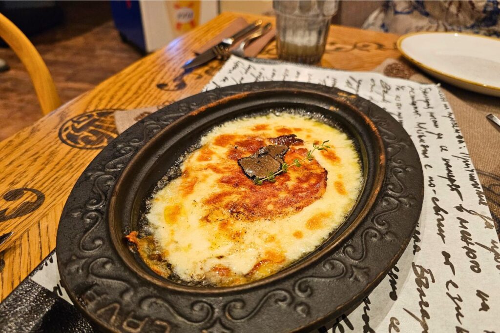 Ein brutzelndes, traditionelles bulgarisches Käsegericht überbacken in einer ovalen Tonschale, garniert mit einer Trüffelscheibe und Thymian im Restaurant Shtastlivetsa.