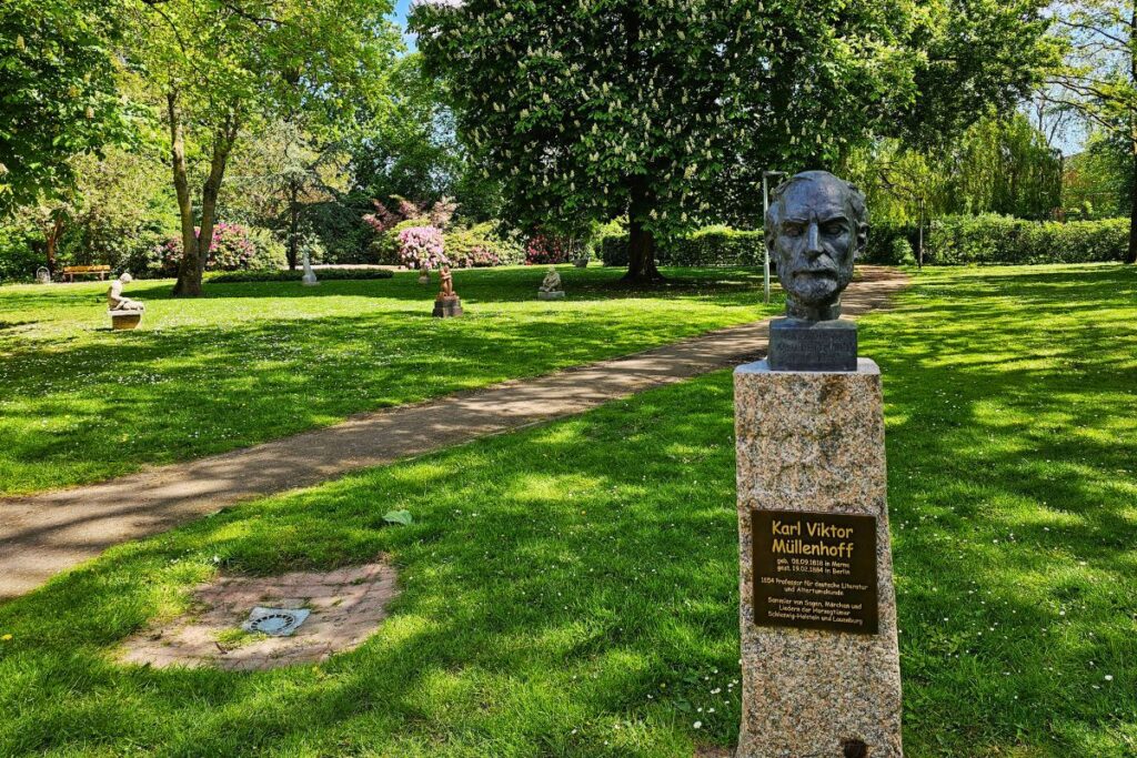 Die Bronzebüste von Professor Karl Viktor Müllenhoff auf einem Steinsockel im grün belaubten Skulpturenpark in Marne unter hohen Bäumen.