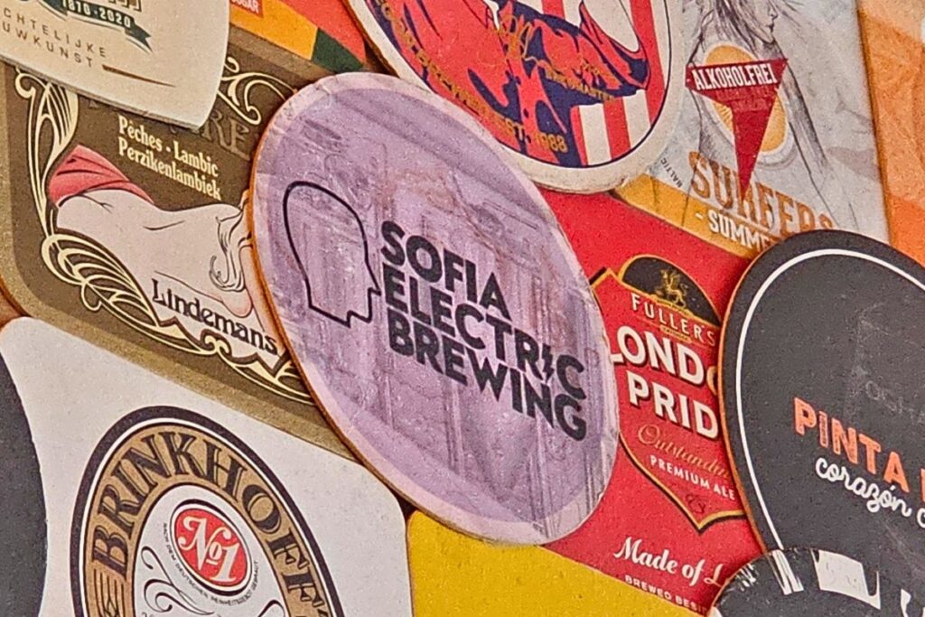 Eine bunte Collage aus verschiedenen Bierdeckeln und Stickern, in deren Mitte ein runder, lila Aufkleber von Sofia Electric Brewing prangt.
