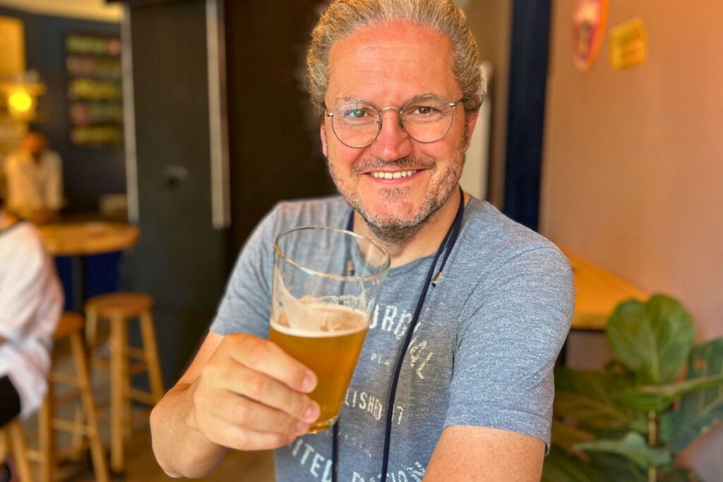 Reiseblogger Nicolo Martin lächelt in einer Bar und hält ein halbleeres Glas helles Craft Beer in der Hand.