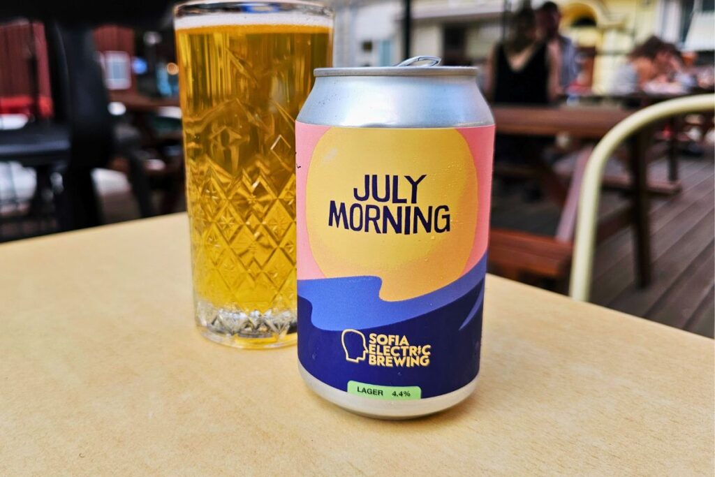 Eine hellbunte Dose „July Morning“ Lagerbier von Sofia Electric Brewing steht neben einem gemusterten Glas hellem Bier auf einem Tisch im Gastgarten.