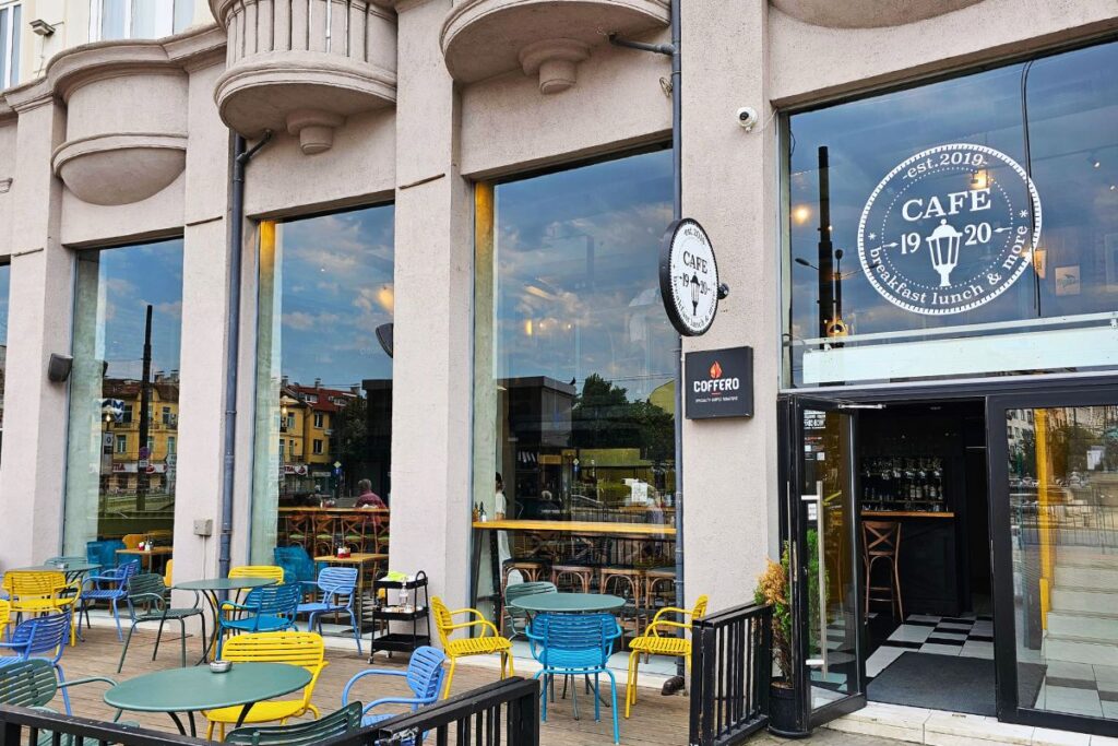 Die stilvolle Außenfassade des Café 1920 in Sofia mit bunten gelben und blauen Stühlen auf der Terrasse vor großen Fensterfronten.