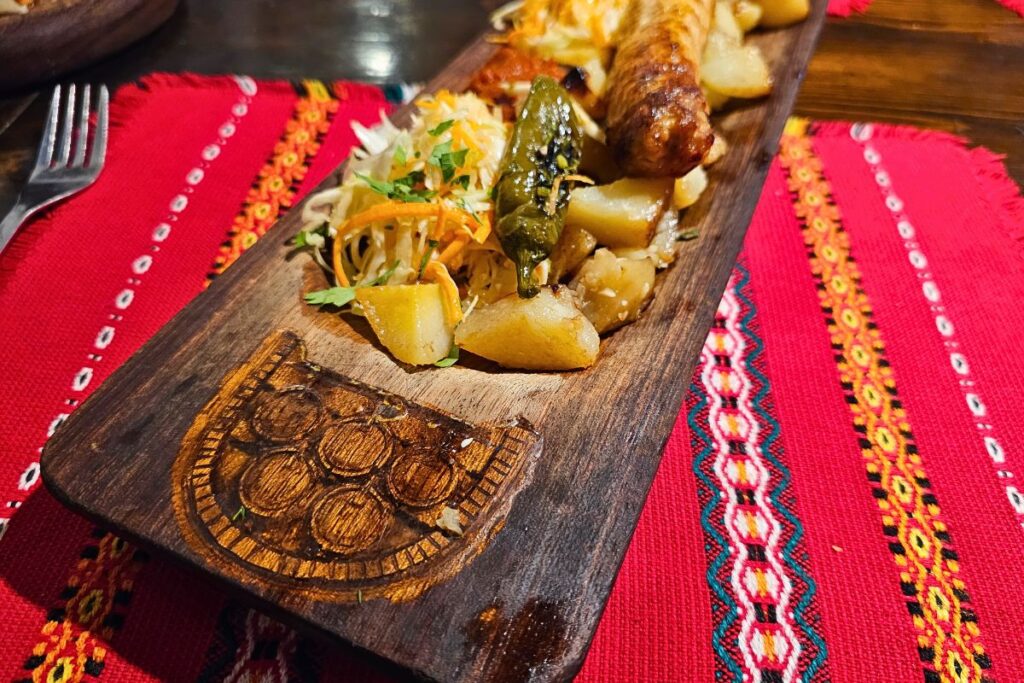 Eine traditionelle bulgarische Grillplatte aus Holz mit Wurst, Kartoffeln und Salat auf einer roten Tischdecke in Hadjidraganov's Cellars in Sofia.
