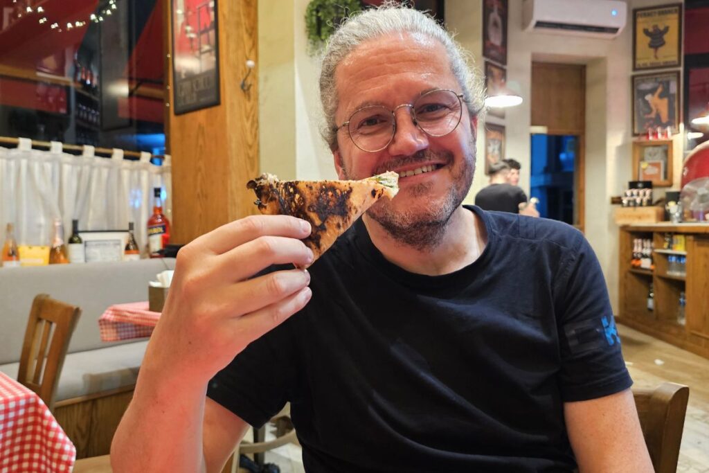 Nicolo Martin hält freudig lächelnd ein frisch gebackenes Stück Pizza in einem gemütlichen Restaurant in Sofia.