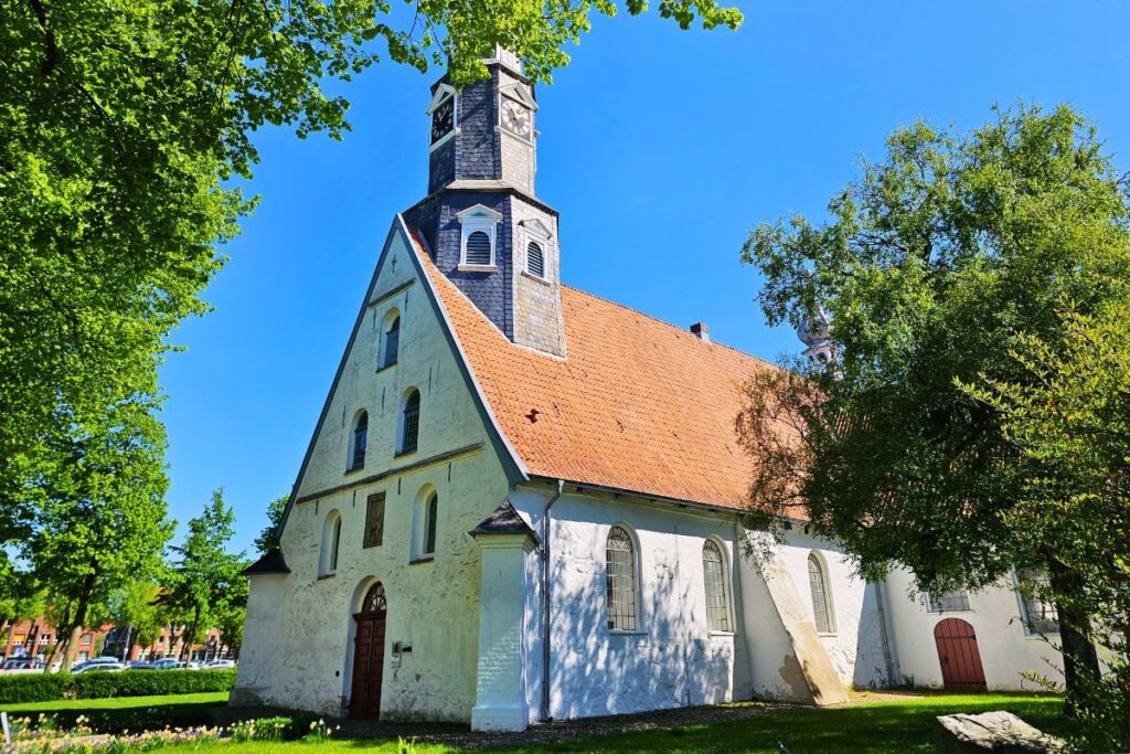 Die weiße Fassade der historischen St. Jürgen Kirche in Heide mit einem hohen, dunklen Turm und Uhrengehäusen vor strahlend blauem Himmel im Sommer.