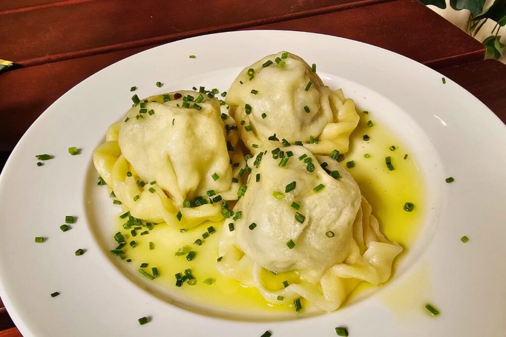 Drei rundlich geformte, meisterhaft gekändelte Kärntner Kasnudeln in warmer Butter mit Schnittlauch auf einem weißen Teller im Restaurant Kärntner-Eck.