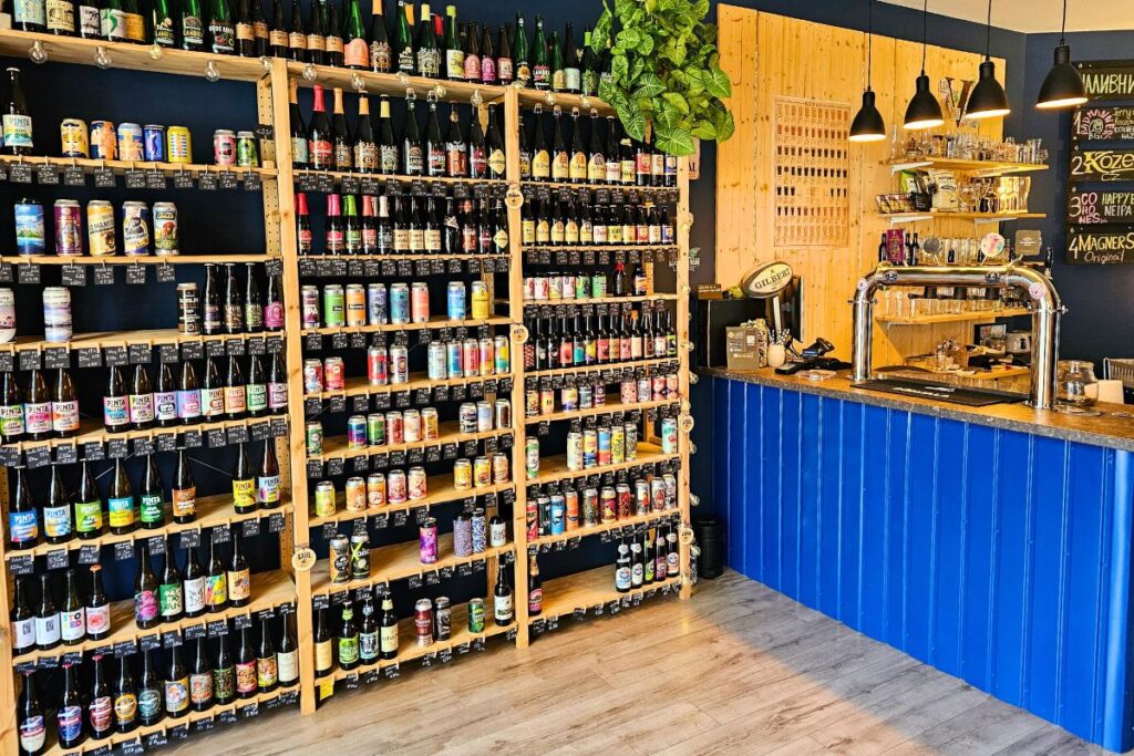 Ein riesiges, bis unter die Decke gefülltes Holzregal mit hunderten Craft-Beer-Flaschen und -Dosen neben einer leuchtend blauen Bar im Vagabond Beer Shop in Sofia.