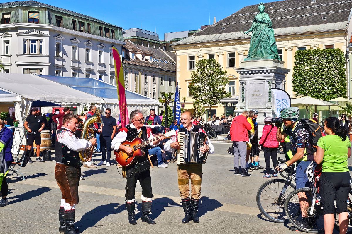 Drei traditionelle Musiker in Kärntner Tracht spielen mit Gitarre, Akkordeon und Baritonhorn auf dem Neuen Platz in Klagenfurt vor dem Maria-Theresia-Denkmal für Radfahrer.