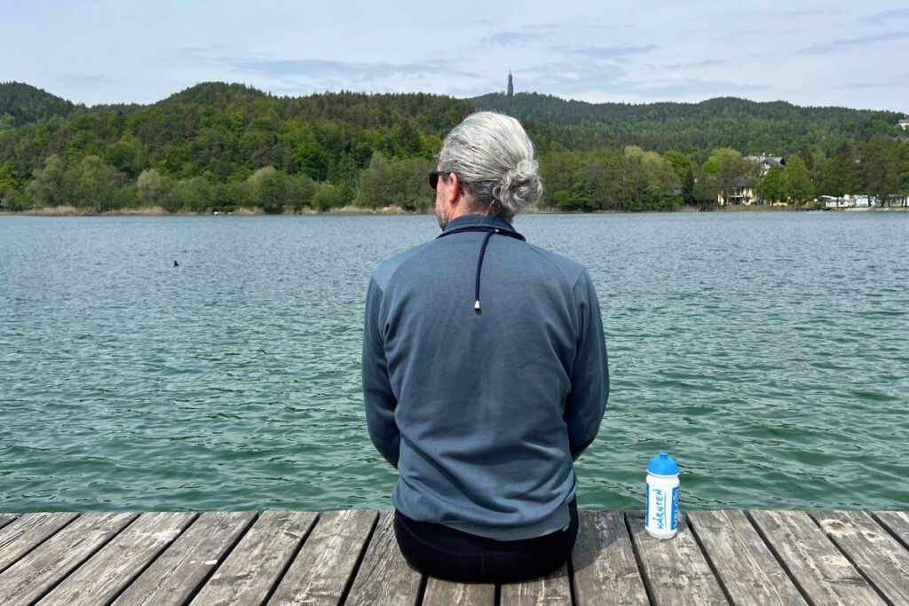 Nicolo Martin sitzt von hinten fotografiert entspannt auf einem Holzsteg am Ufer des Keutschacher Sees, neben ihm liegt eine Sporttrinkflasche.
