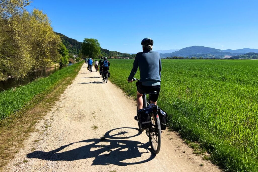 Blick von hinten auf Nicolo Martin auf einem hellen Schotterweg, rechts ein weites, saftig grünes Feld unter strahlend blauem Himmel und eine Gruppe Radler voraus.