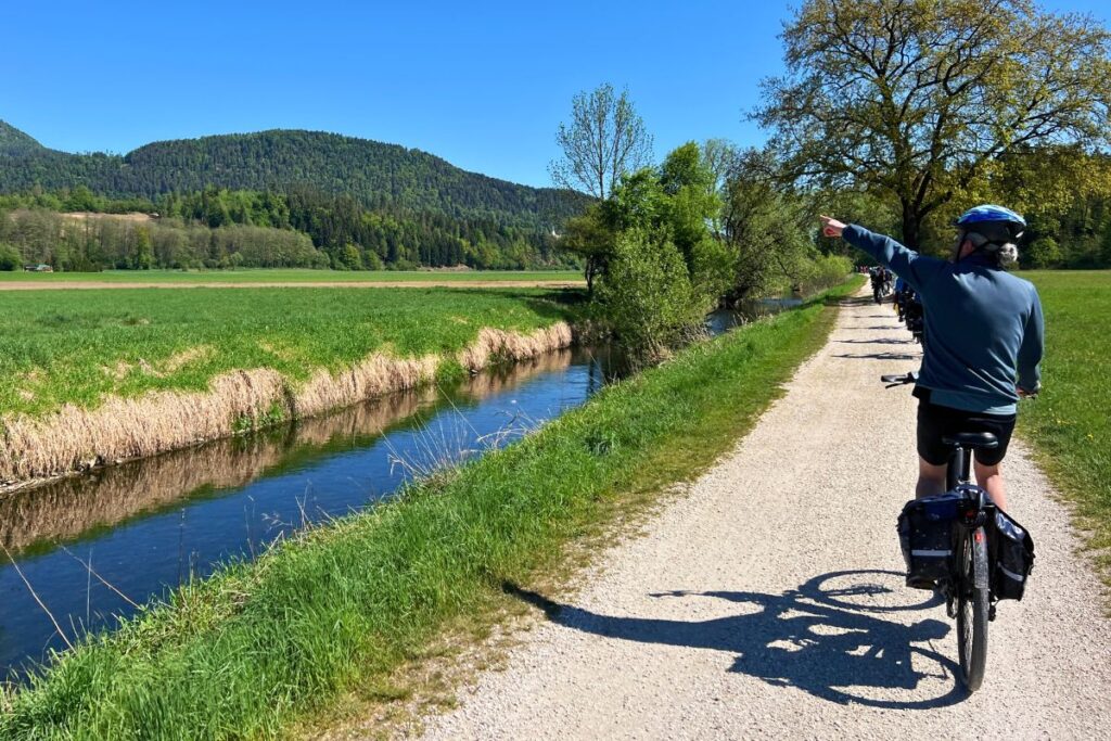 Nicolo Martin zeigt von hinten fotografiert auf einem Schotterweg mit ausgestrecktem Arm nach vorne rechts, links fließt ein schmaler Kanal parallel zum Weg.