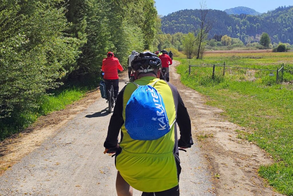 Blick von hinten auf einen Radfahrer in gelb-schwarzem Trikot mit einem blauen Kärnten-Rucksack auf einem Schotterweg, gefolgt von weiteren Radlern entlang von Bäumen und Feldern.